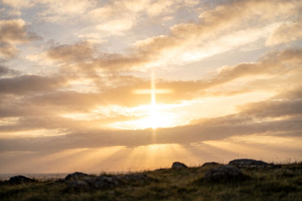 Sunset sky with a glowing cross-shaped beam of light shining through clouds above a rocky, grassy landscape